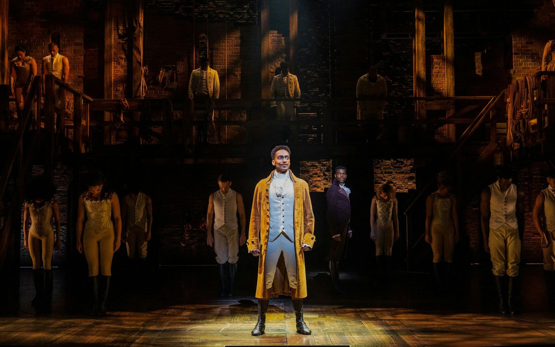 Cast performing on stage in Hamilton production, featuring lead actor in period costume