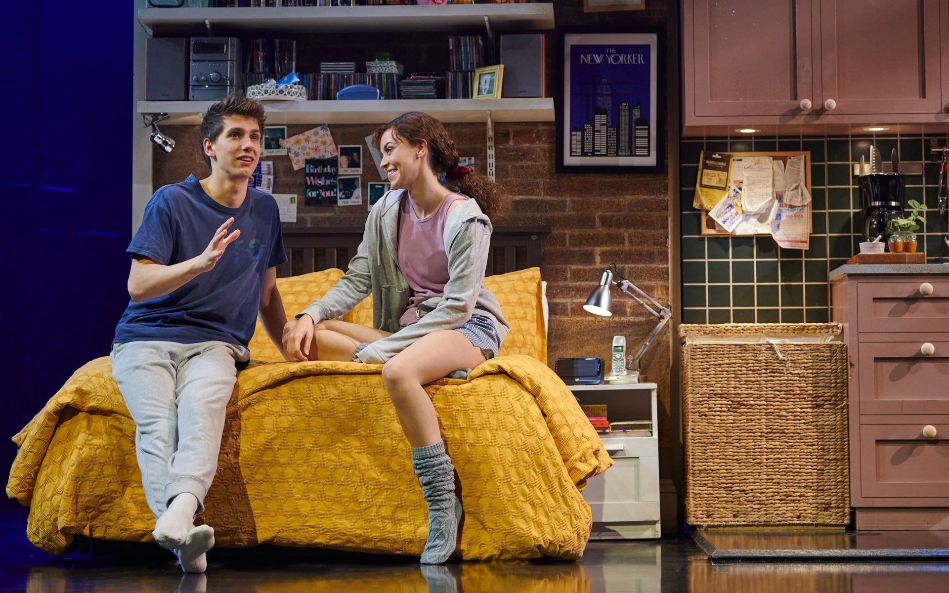 Actors on stage during The Devil Wears Prada West End show, sitting on a bed in a cozy room