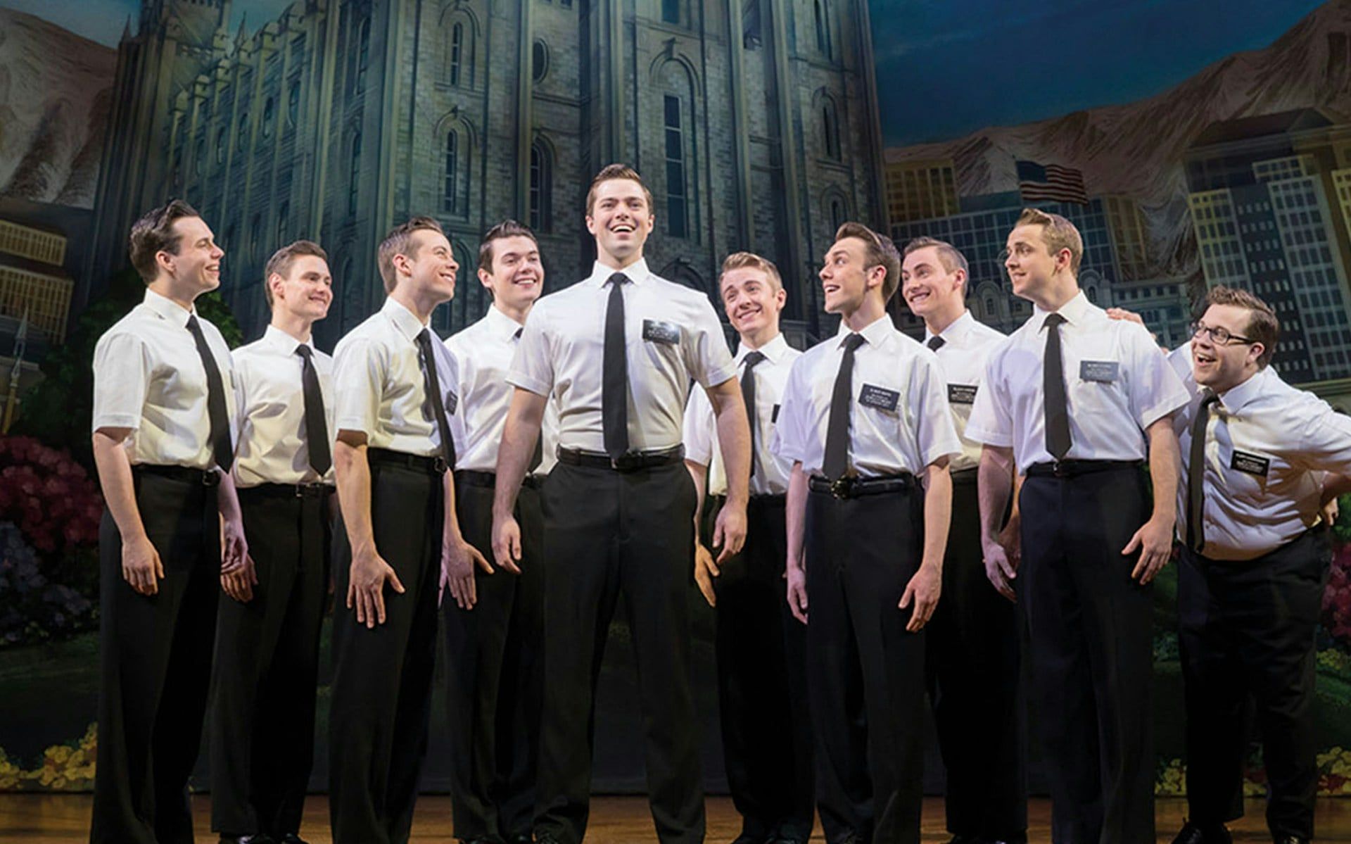 Cast of "The Book of Mormon" performing on stage with a backdrop of a cityscape