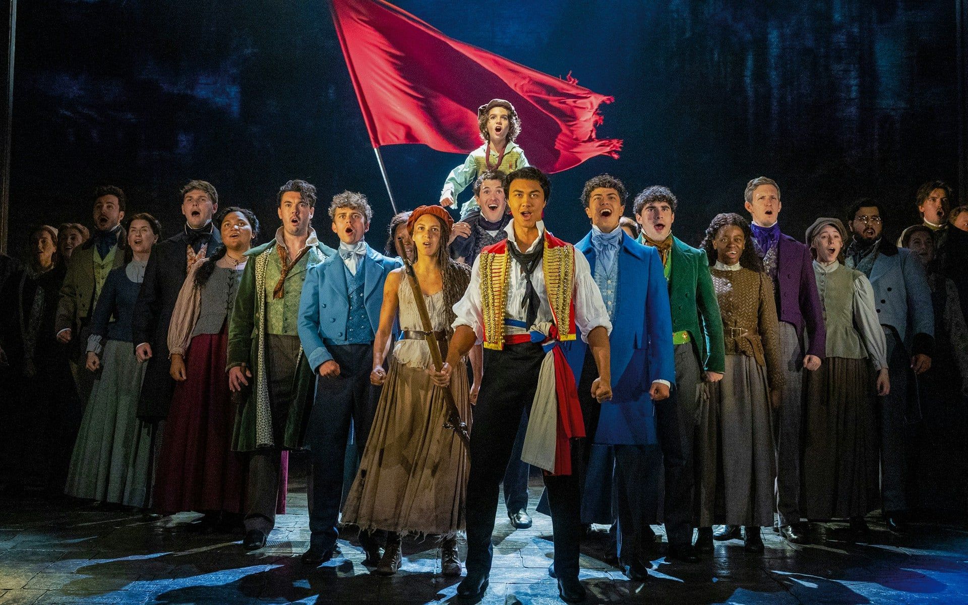 Les Misérables cast performing on stage with a red flag in the background