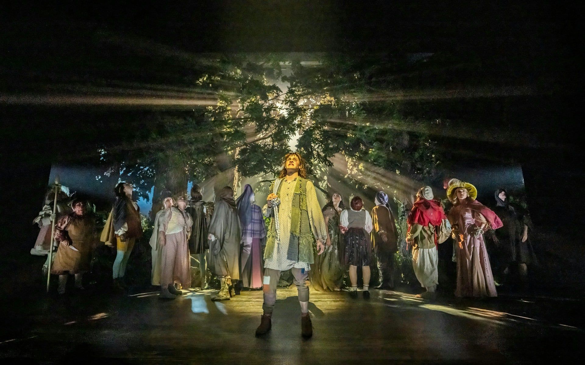 Cast performing in "Into The Woods" with forest backdrop and dramatic lighting