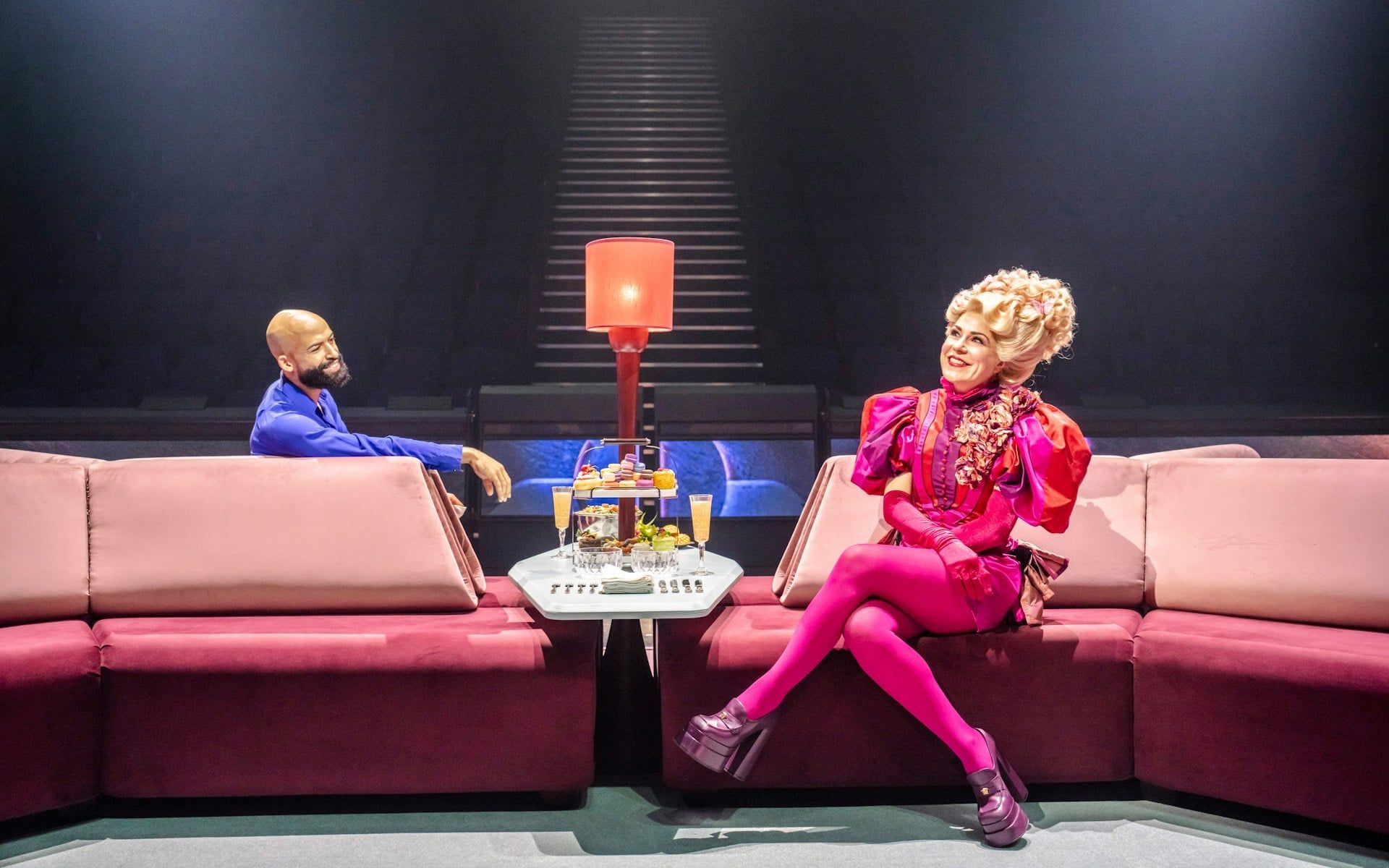 Stage scene from Hunger Games West End show with two actors seated on a pink sofa — The Hunger Games