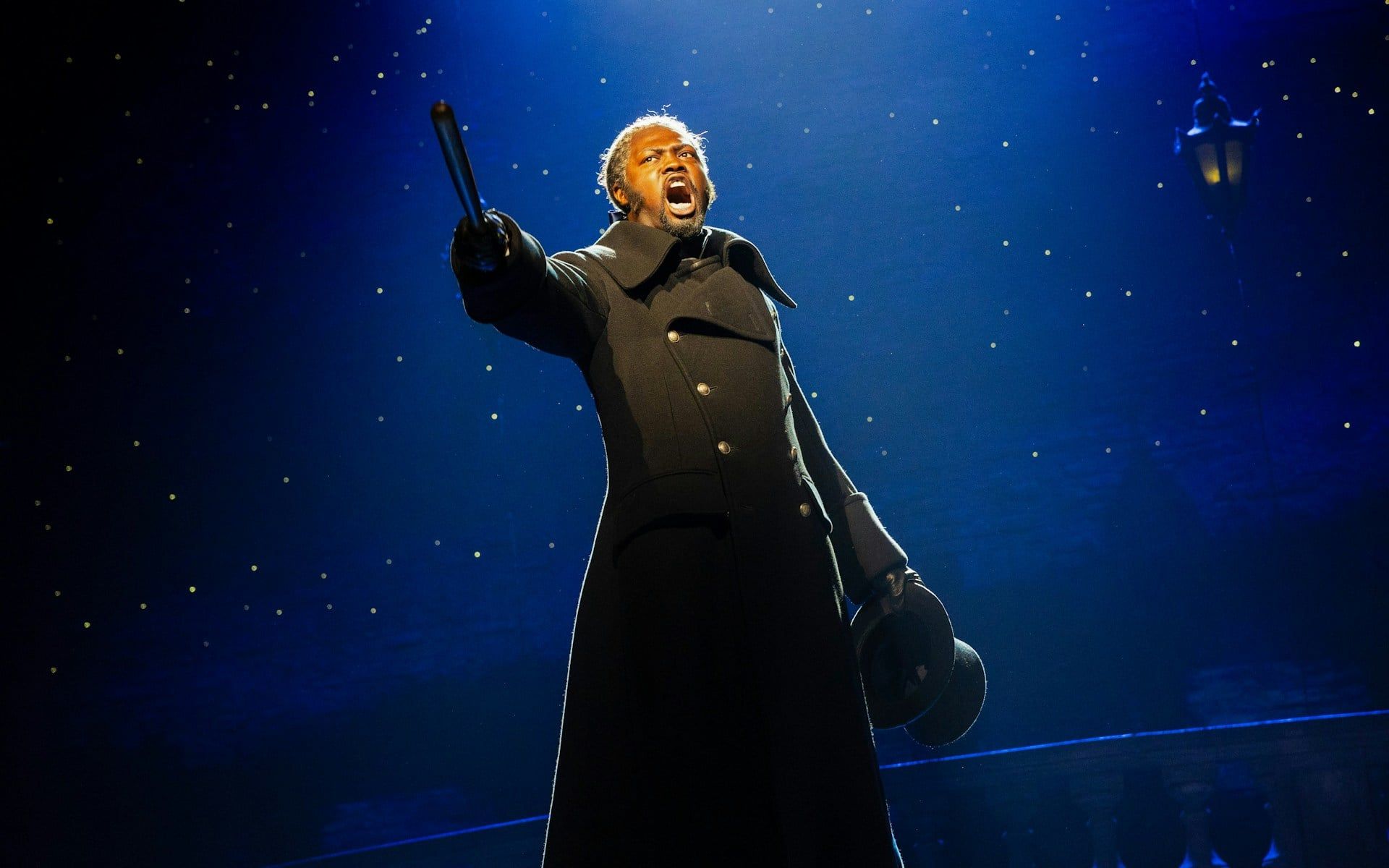 Performer on stage during Les Misérables musical, holding a hat and pointing dramatically