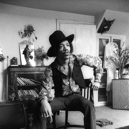A man wearing a hat and patterned shirt sits on a chair in a cluttered room with a cabinet, books, and a television.
