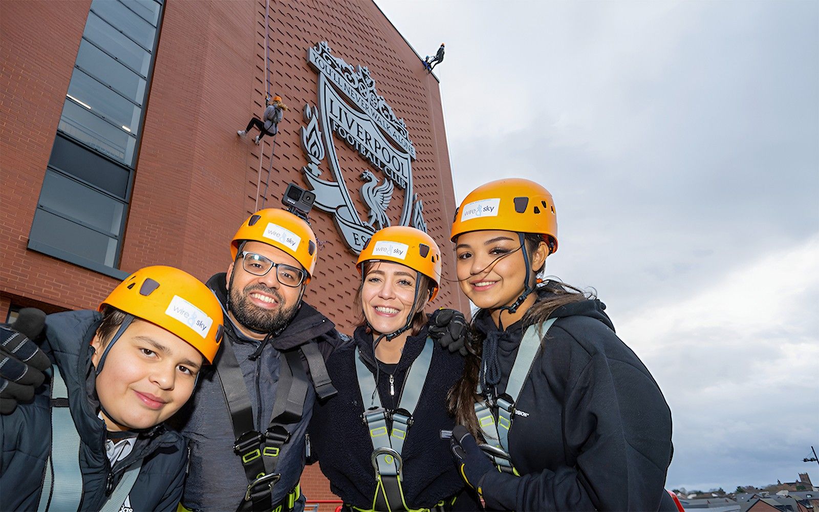 Group preparing for abseil at Liverpool FC's Anfield Stadium with club emblem in background.