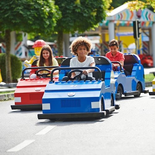 Three children drive mini cars on a small road in an amusement park. The cars are blue and red, and trees are in the background.