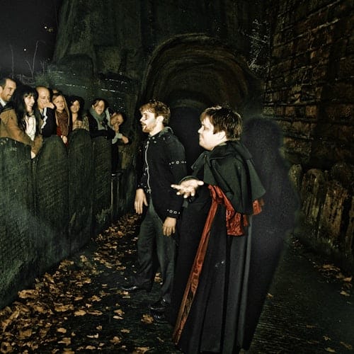 Two costumed performers interact with a group of onlookers by an old, stone wall covered in fallen leaves at night.