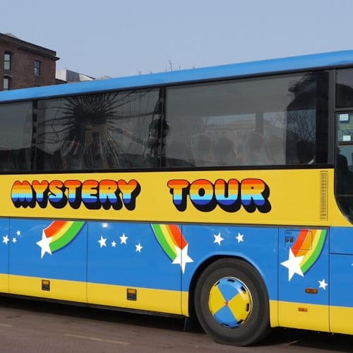 A colorful bus with "Mystery Tour" written on its side, adorned with rainbows and stars, parked in an urban area.