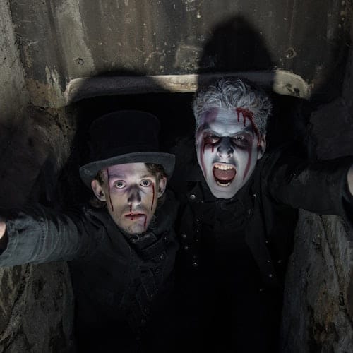 Two people with dramatic makeup and fake blood look upwards from a dark, enclosed space; one wears a top hat.